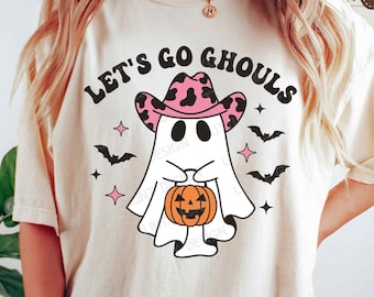 May include: Cream-colored t-shirt featuring a cartoon ghost wearing a pink cowboy hat and holding a jack-o'-lantern. The text "LET'S GO GHOULS" is arched above the ghost. Bats and stars surround the ghost, creating a Halloween theme.