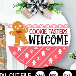 May include: A pink and white round wooden sign with the text "Cookie Tasters Welcome" in black. A gingerbread man cookie graphic is on the left side of the sign. The sign is decorated with white snowflakes and gingerbread men.
