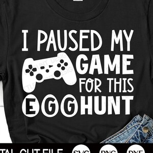 Op de afbeelding: Zwart T-shirt met witte tekst die "Ik heb mijn game gepauzeerd voor deze eierenjacht" zegt. Boven de tekst staat een witte afbeelding van een videogamecontroller.