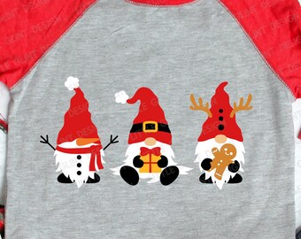 Könnte beinhalten: Ein graues und rotes Raglan-T-Shirt mit einem Design aus drei Gnomen. Die Gnome tragen rote Mützen und haben weiße Bärte. Ein Gnom hält ein Lebkuchenmännchen, ein anderer ein Geschenk und der dritte einen Schneemann.