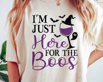 May include: A white t-shirt with the text "I'm Just Here For The Boo's" in black and purple lettering. A purple wine glass with a witch's hat on top and bats flying around it are also on the shirt.