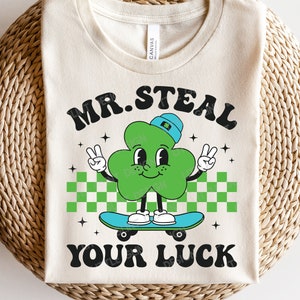 Pode incluir: Uma camiseta branca com um personagem de trevo verde em um skate. O trevo está usando um chapéu azul e tem um rosto sorridente. O texto "MR. STEAL YOUR LUCK" está impresso na camiseta.