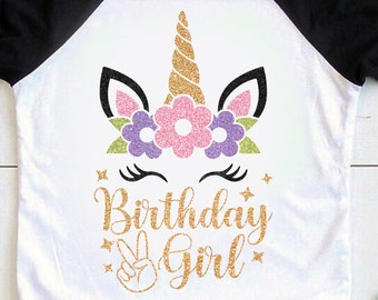 May include: A white t-shirt with a black and white baseball style sleeve design. The shirt features a gold glitter unicorn with pink, purple, and green flowers. The text "Birthday Girl" is written in gold glitter with a peace sign.