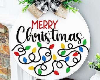 May include: A white, round Christmas sign with the words "MERRY Christmas" in red and black. A string of colorful Christmas lights is below the text. The sign is decorated with a buffalo plaid bow and greenery.