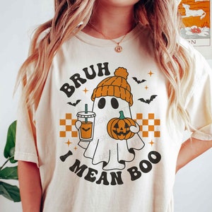 May include: A white t-shirt with a ghost wearing a pumpkin hat and holding a pumpkin and a cup of coffee. The text on the shirt says "Bruh I mean Boo".