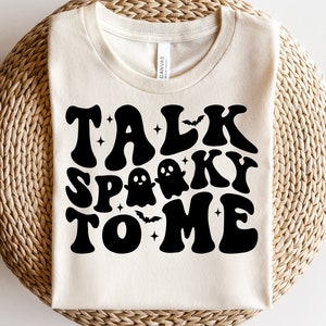 May include: A white t-shirt with the text "TALK SPOOKY TO ME" in black retro-style lettering. The text is decorated with stars and a ghost.