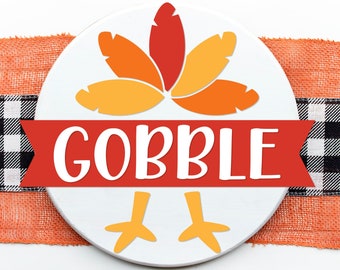 May include: A white circle with a red banner that says "GOBBLE" and a turkey design with orange, yellow, and red feathers. The circle is surrounded by a black and white checkered ribbon and an orange burlap ribbon.