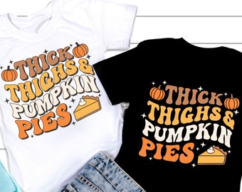 Könnte beinhalten: Zwei T-Shirts mit einem grafischen Design mit dem Text "Thick Thighs & Pumpkin Pies" in einer Retro-Schriftart. Das Design enthält auch einen Kürbis und ein Stück Kürbiskuchen.