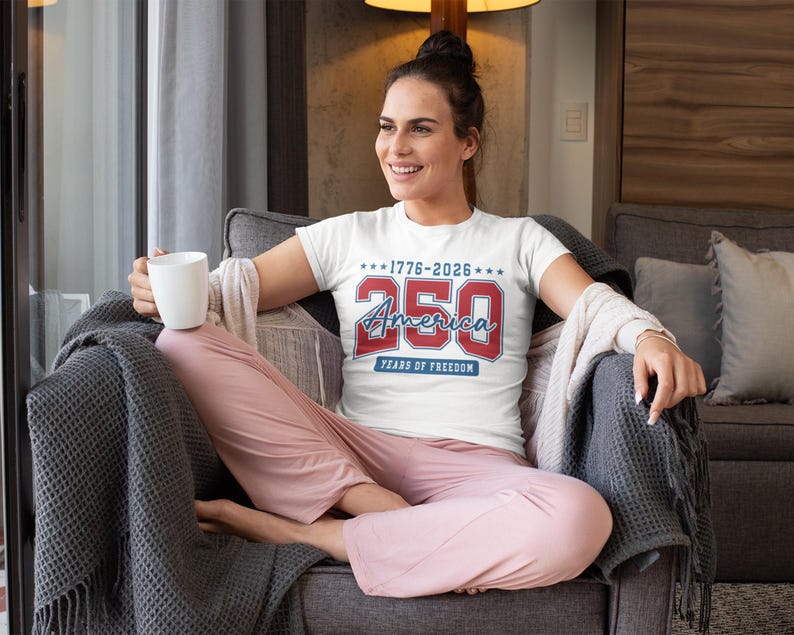 May include: White t-shirt with the text "1776-2026 250 America Years of Freedom" in red and blue. The shirt is worn by a person sitting in a chair, wearing pink pants and holding a white mug.