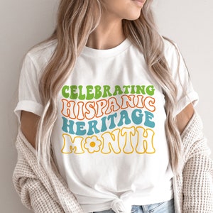 May include: White t-shirt with a colorful graphic that says "Celebrating Hispanic Heritage Month" in a retro font.