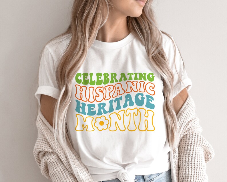 May include: White t-shirt with a colorful graphic that says "Celebrating Hispanic Heritage Month" in a retro font.