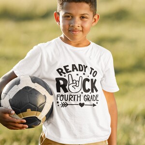May include: A white t-shirt with a black graphic that says "Ready to Rock Fourth Grade" with a rock and roll hand gesture and an arrow with a heart. A young person is wearing the shirt and holding a soccer ball.