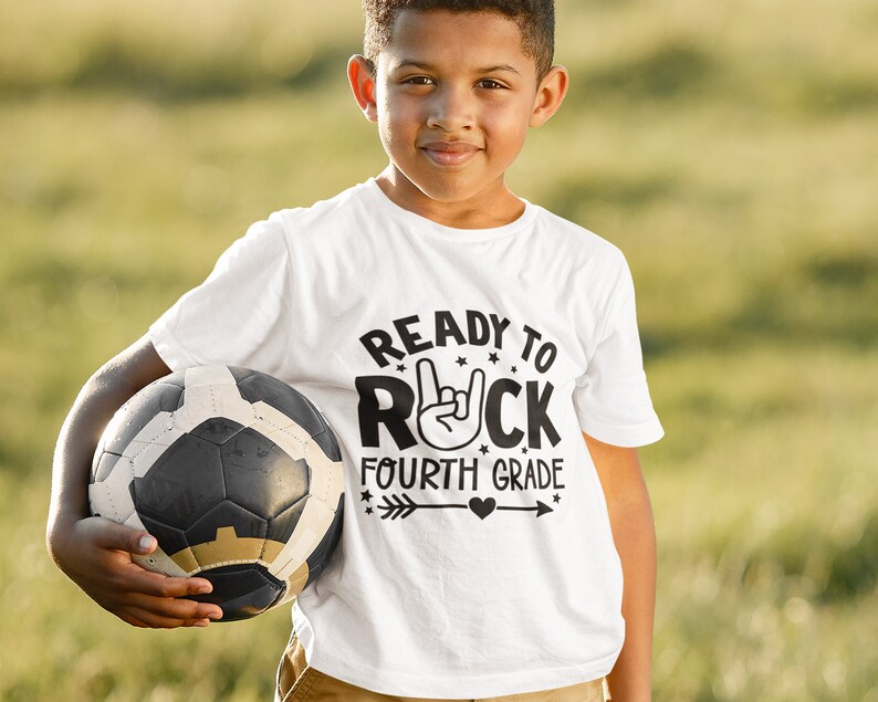 May include: A white t-shirt with a black graphic that says "Ready to Rock Fourth Grade" with a rock and roll hand gesture and an arrow with a heart. A young person is wearing the shirt and holding a soccer ball.