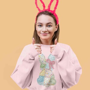 May include: A light pink sweatshirt featuring a patchwork bunny design with floral and gingham patterns. The model is wearing a pink bunny ear headband. The background is a solid light orange color.