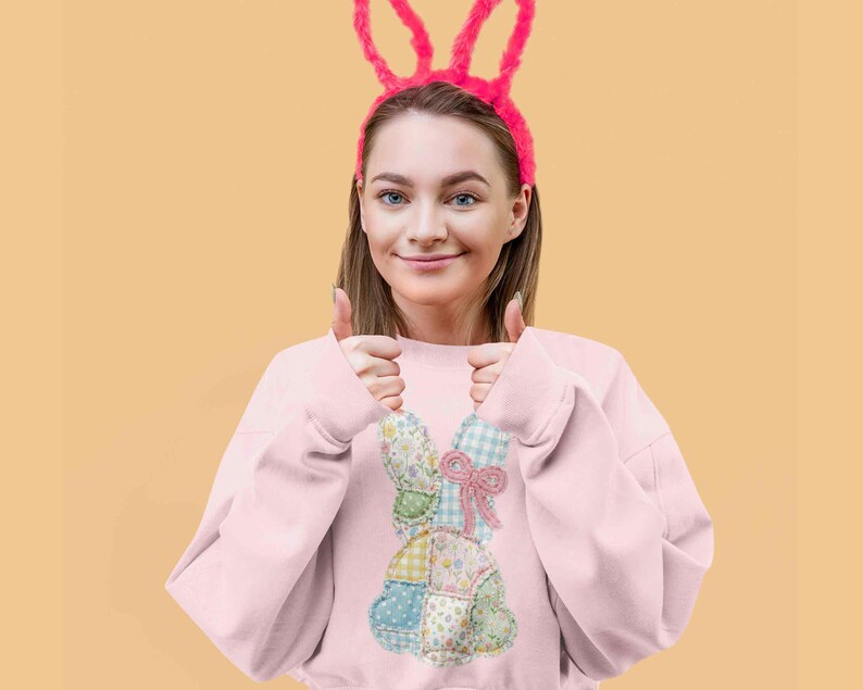 May include: A light pink sweatshirt featuring a patchwork bunny design with floral and gingham patterns. The model is wearing a pink bunny ear headband. The background is a solid light orange color.