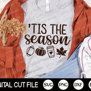 May include: A light grey t-shirt featuring the text "'Tis the Season" in brown script, with a football, pumpkin, coffee cup, and maple leaf design. The shirt is styled with a rust-coloured jacket, jeans, and white trainers.
