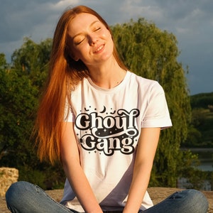 May include: A woman wearing a white t-shirt with a black graphic that says "Ghoul Gang" with a crescent moon, stars, and bats. She is sitting on a rock with her legs crossed and her eyes closed.