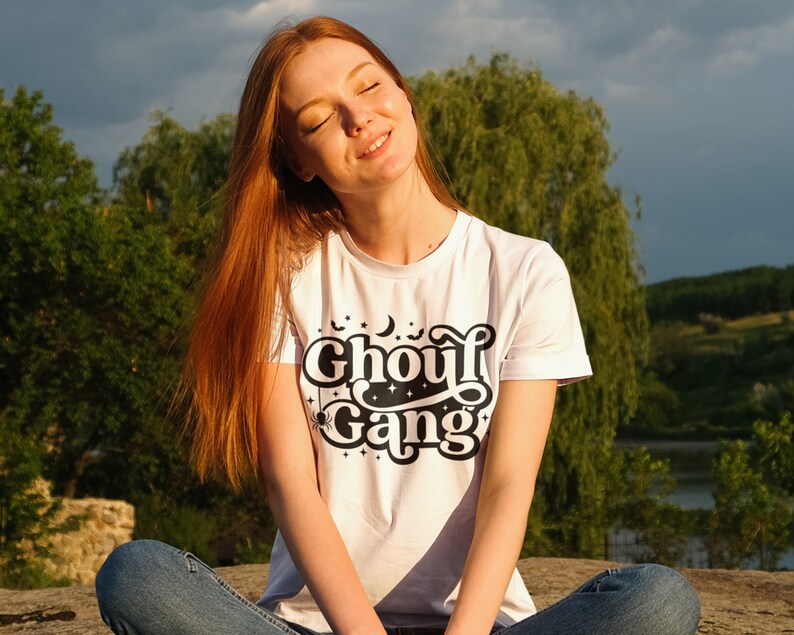 May include: A woman wearing a white t-shirt with a black graphic that says "Ghoul Gang" with a crescent moon, stars, and bats. She is sitting on a rock with her legs crossed and her eyes closed.