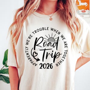 Pu&ograve; includere: T-shirt color crema con grafica nera che recita "Road Trip 2026". Il design include un sole, una freccia e la frase "Apparently We're Trouble When We Are Together" disposta a cerchio attorno al testo centrale.