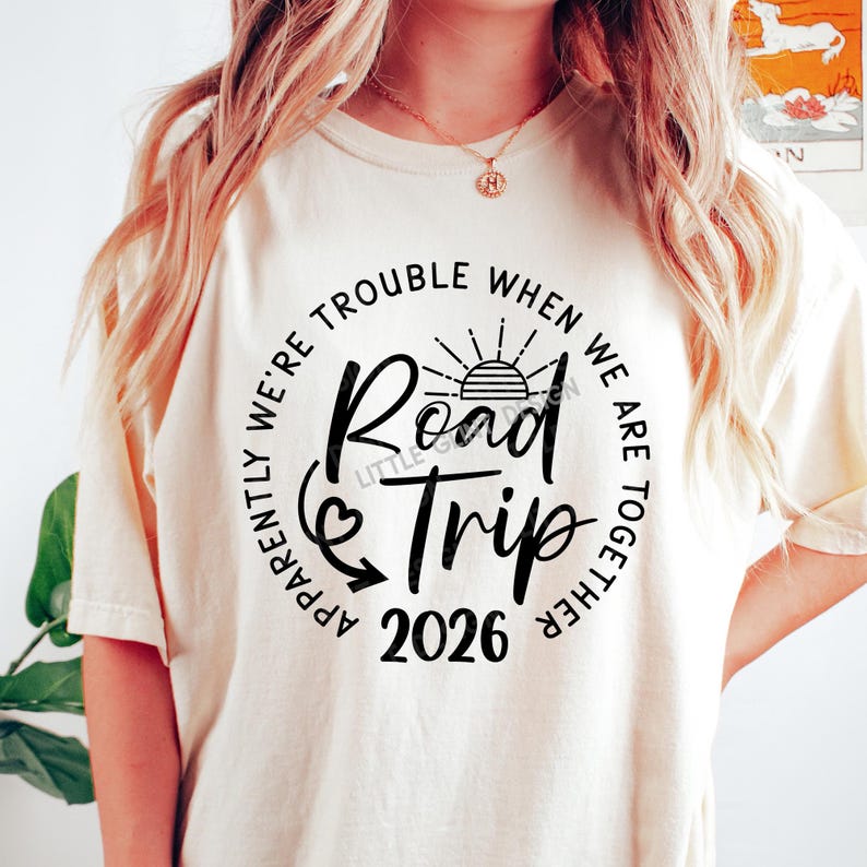 Pu&ograve; includere: T-shirt color crema con grafica nera che recita "Road Trip 2026". Il design include un sole, una freccia e la frase "Apparently We're Trouble When We Are Together" disposta a cerchio attorno al testo centrale.