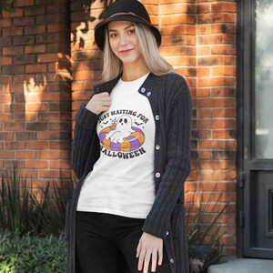 May include: White t-shirt with the text "Just Waiting for Halloween" and a cartoon ghost floating on a pool float. The ghost is white with black eyes and is wearing a drink. The float is orange, purple, and white.
