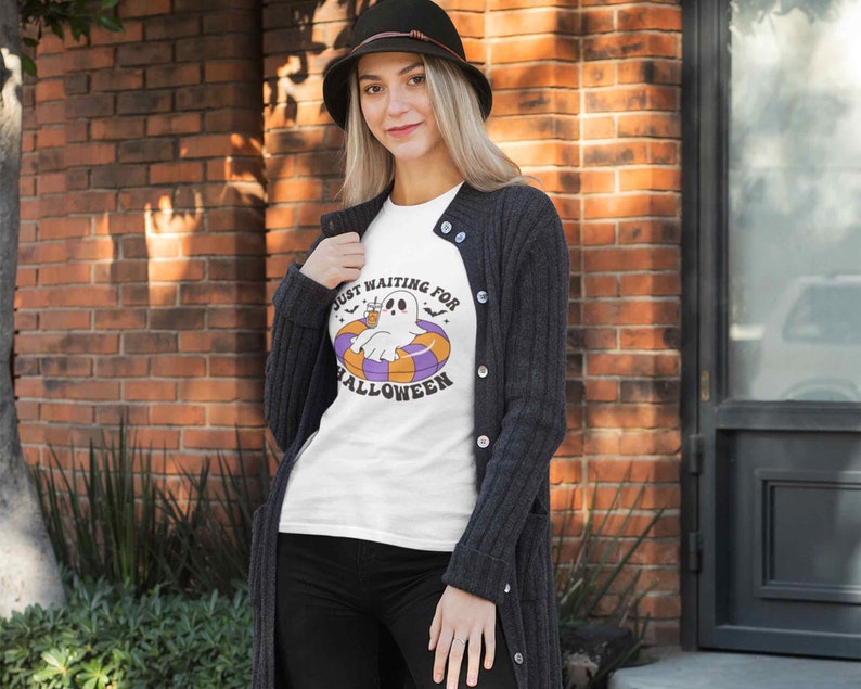 May include: White t-shirt with the text "Just Waiting for Halloween" and a cartoon ghost floating on a pool float. The ghost is white with black eyes and is wearing a drink. The float is orange, purple, and white.
