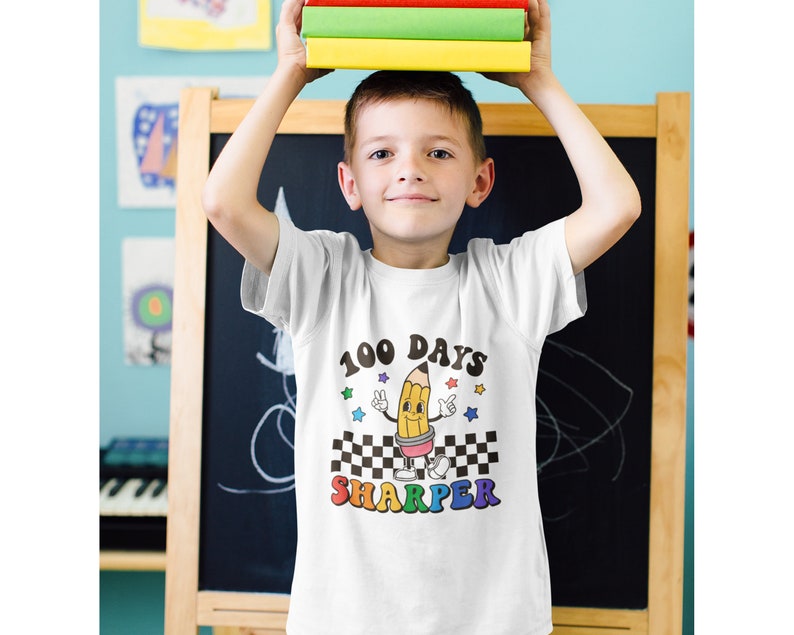May include: A white t-shirt with a yellow pencil graphic and the text "100 Days Sharper". The pencil has a smiling face and is holding up two fingers in a peace sign. There are rainbow stars around the pencil.