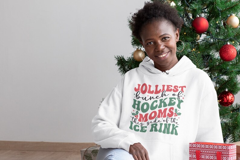 May include: A white hoodie with the text "Jolliest bunch of Hockey Moms this side of the Ice Rink" in red, green, and white. The image features a woman smiling in front of a Christmas tree.
