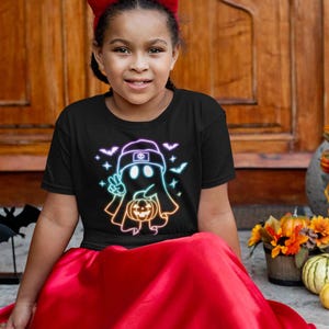 May include: A young person wearing a black t-shirt with a neon rainbow ghost holding a jack-o-lantern. The ghost is wearing a backwards baseball cap. The t-shirt is a Halloween themed design.