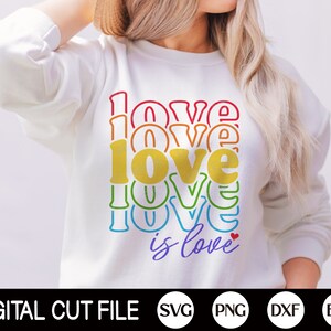 Puede incluir: Una sudadera blanca con un gr&aacute;fico de colores arco&iacute;ris que dice "love is love" con un peque&ntilde;o coraz&oacute;n rojo.
