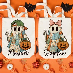 May include: Two white Halloween tote bags with cream-colored handles. Each bag features a ghost character with a patchwork design, holding a jack-o'-lantern. One ghost wears a green cap, the other a pink bow. The names "Mason" and "Sophia" are written below.