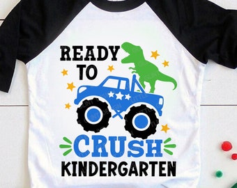 Puede incluir: Una camiseta blanca con un dinosaurio negro y verde en un camión monstruo azul. El texto "Ready to Crush Kindergarten" está impreso en la camiseta.