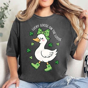 May include: Dark gray t-shirt featuring a white goose with a green bow, boots, and shamrocks. The text "LUCKY GOOSE ON THE LOOSE" is printed above the goose. The shirt is a casual style.