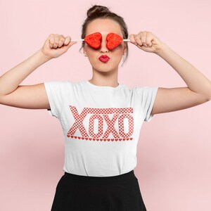Pu&ograve; includere: Una maglietta bianca con una grafica "XOXO" a scacchi rossi e bianchi. La maglietta ha una fila di piccoli cuori rossi sotto la grafica.