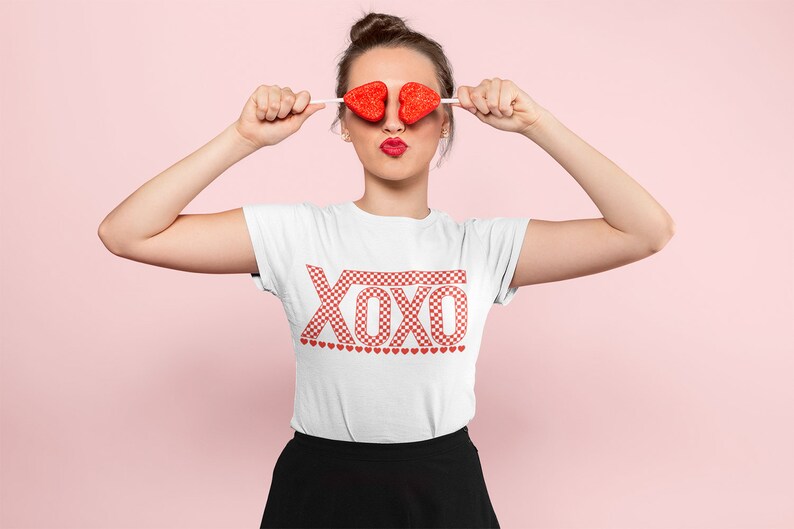 Pu&ograve; includere: Una maglietta bianca con una grafica "XOXO" a scacchi rossi e bianchi. La maglietta ha una fila di piccoli cuori rossi sotto la grafica.