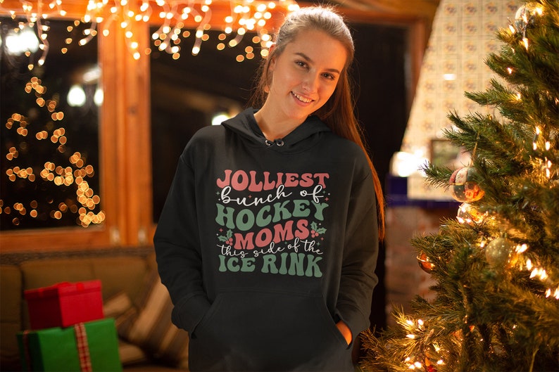 May include: Black hoodie with the text "Jolliest bunch of Hockey Moms this side of the Ice Rink" in pink, green, and white. The hoodie has a drawstring hood and a front pocket. The background includes a Christmas tree and string lights.