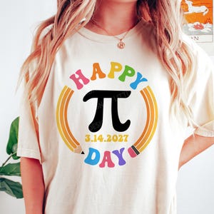 Może przedstawiać: Kremowa koszulka z kolorową grafiką na Dzień Liczby Pi. Projekt zawiera słowa "HAPPY DAY" w tęczowych kolorach, symbol Pi, datę 3.14.2027 oraz obramowanie oł&oacute;wkiem.