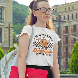 May include: White t-shirt with a graphic of a smiling pumpkin holding a pencil and the text "I teach the cutest Pumpkins in the patch".