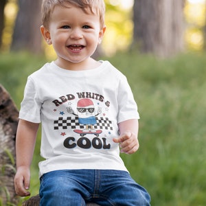 May include: A white t-shirt with a red, white, and blue cartoon character skateboarding. The character is wearing sunglasses and has a peace sign on each hand. The text on the shirt reads "Red White & Cool".