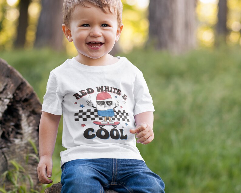 May include: A white t-shirt with a red, white, and blue cartoon character skateboarding. The character is wearing sunglasses and has a peace sign on each hand. The text on the shirt reads "Red White & Cool".