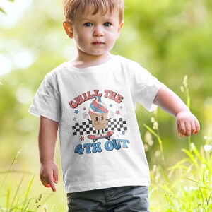 May include: White t-shirt with a graphic of an ice cream cone on a skateboard. The ice cream cone is wearing a red, white, and blue hat and has a peace sign. The text on the shirt says "Chill the 4th Out".