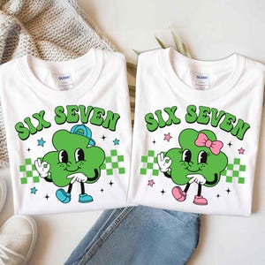 May include: Two white t-shirts with the text "SIX SEVEN" in green. Each shirt features a cartoon clover with a hat or bow, hands making an "okay" sign, and checkered green and white accents. Blue jeans and a pair of white sneakers are in the image.