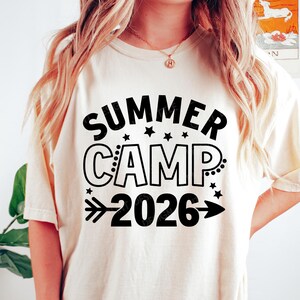 May include: Cream-colored t-shirt with the words "SUMMER CAMP 2026" in black, with stars and an arrow design. The shirt is made of a soft material and is suitable for summer camp activities.