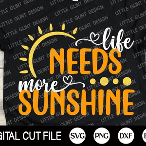 Op de afbeelding: Een zwart T-shirt met een oranje zon, witte tekst die "life needs more sunshine" luidt en een kleine hartgrafiek. De tekst is in een handschrift.