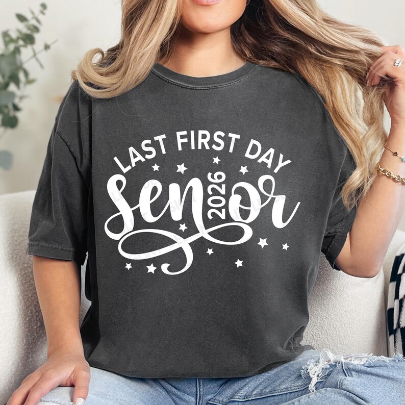 May include: Dark gray t-shirt with the text "LAST FIRST DAY SENIOR 2026" in white script and stars. The shirt is a casual style, suitable for everyday wear. The design is centered on the front.