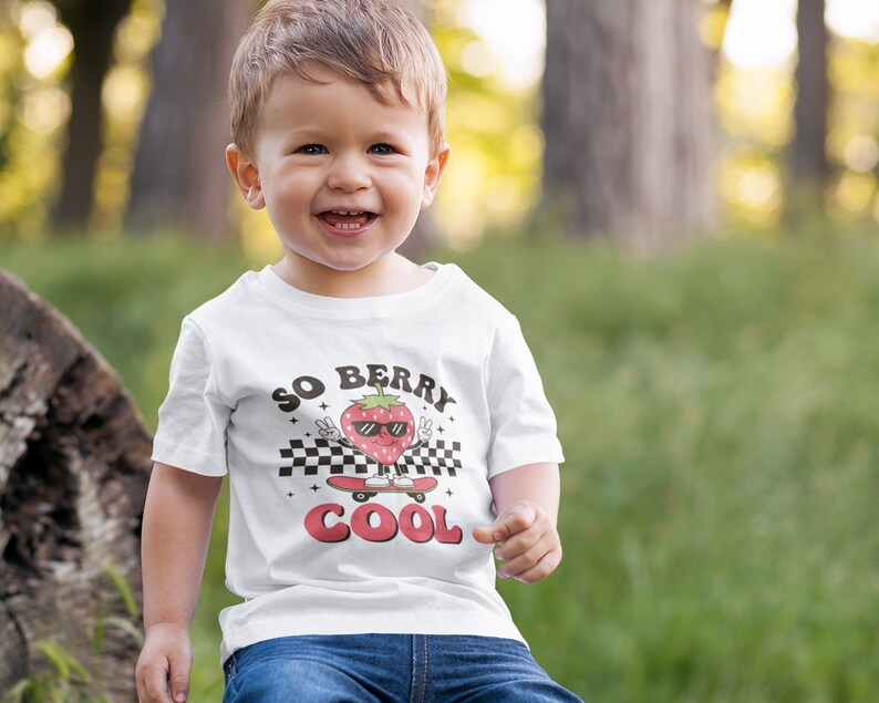 K&ouml;nnte beinhalten: Ein wei&szlig;es T-Shirt mit einem Aufdruck einer Erdbeere, die eine Sonnenbrille tr&auml;gt und Skateboard f&auml;hrt. Der Text "SO BERRY COOL" ist auf dem Shirt aufgedruckt.