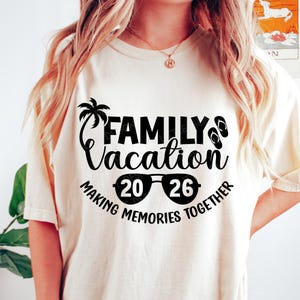 May include: Cream-colored t-shirt with a black graphic that reads "FAMILY Vacation 20 26 MAKING MEMORIES TOGETHER." The design includes a palm tree, sunglasses, and flip-flops. The shirt is a casual wear item.