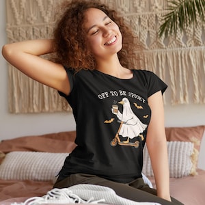 May include: Black t-shirt with the text "OFF TO BE SPOOKY" and a cartoon ghost riding a scooter while holding a coffee cup. The ghost is wearing a white sheet and is accompanied by bats. The shirt is a Halloween-themed design.