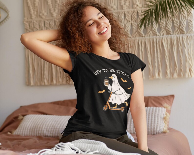 May include: Black t-shirt with the text "OFF TO BE SPOOKY" and a cartoon ghost riding a scooter while holding a coffee cup. The ghost is wearing a white sheet and is accompanied by bats. The shirt is a Halloween-themed design.
