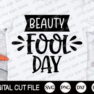 Op de afbeelding: Een wit T-shirt met zwarte tekst die "Beauty Fool Day" luidt. De tekst is in een gestileerd lettertype met een decoratieve versiering. Het shirt ligt plat op een houten oppervlak met een wortel en een roze ei op de achtergrond.